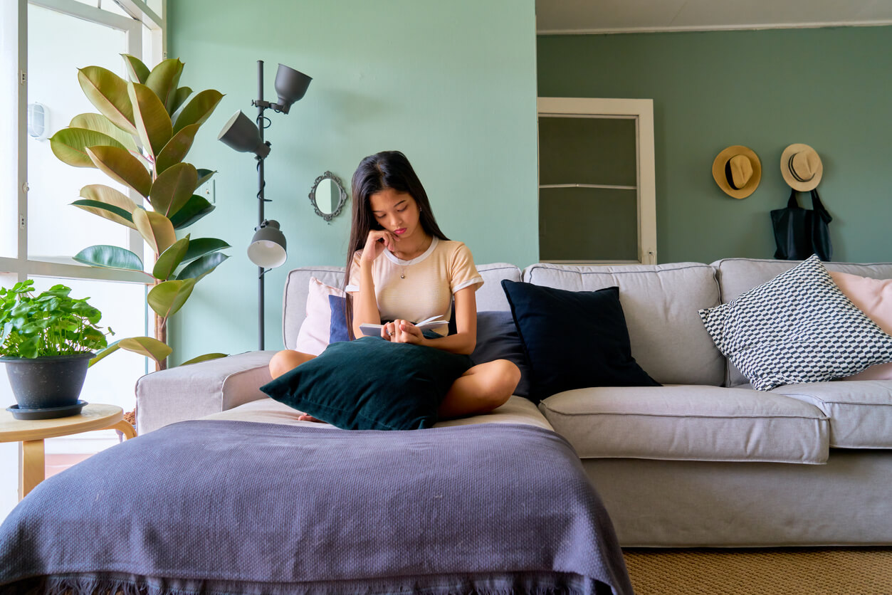 Jeune étudiante asiatique assise dans le salon de son appartement étudiant Paris 13. Elle lit un livre et l'on peut voir la beauté de son appartement, avec un canapé, des plantes vertes, une grande fenêtre vitrée et un mur vert pastel