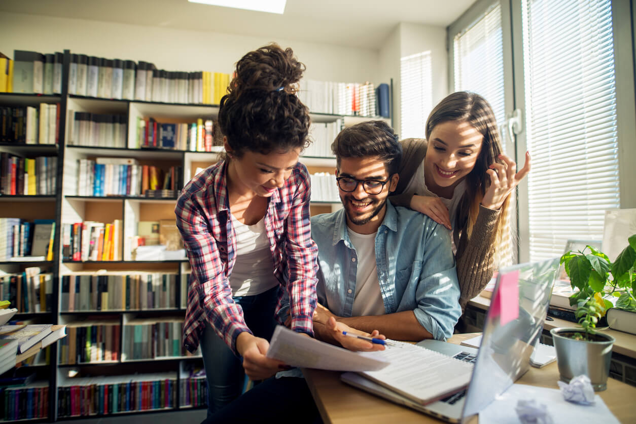 Un étudiant assis au milieu de deux étudiantes, en train de travailler joyeusement ensemble dans une salle d'études. On voit des livres posés dans une étagère derrière eux, un ordinateur portable posé sur la table devant eux. Ils regardent des feuilles de papier. Cette image montre le partage et la convivialité dont bénéficient les étudiants en location étudiant Paris